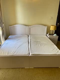 2 white single beds with 1 side table along with mattress.